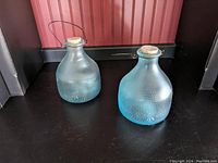 Pair of decorative glass jars with blue gradient tint and cork lids on black surface against a reddish background.