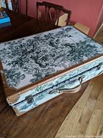Photo showing the top and sides of the suitcase covered in gray and green toile fabric with leather trim and brass hardware