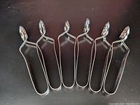 Set of six shiny stainless steel escargot tongs arranged side by side on a dark surface.