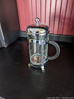 French press coffee or tea maker showing glass carafe, metal lid with knob, metal frame, and black handle on black surface.