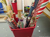 Photo showing red plastic container filled with assorted rolls of holiday wrapping paper with visible designs including stripes, Santa Claus, and text.