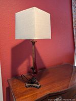 Photo of side view of candlestick-style lamp showing the beige square lampshade, dark metal base and pole with tropical leaf motifs, and red tassel.