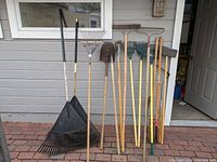 Photo showing wide plastic leaf rake, metal fan-shaped rake with wooden handle, curved rake, shovel and other tools in a line against a wall.