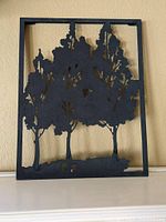 Frontal view of black metal art with tree silhouettes within a rectangular frame placed against a beige wall.