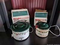 Two Rival mini crock pots with original boxes behind them on a black surface, one larger with grapevine design, the other smaller with fruit design