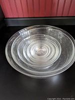 Stacked set of 9 clear nesting mixing bowls on a black surface, showing size gradation and concentric circles detail inside bowl surfaces.