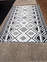 Full view of a rectangular outdoor rug laid on a concrete surface, showing the entire black and white geometric pattern design.