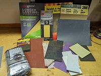 Photo showing a variety of sandpaper sheets, sanding sponges, sanding blocks, and steel wool pack all arranged on surface