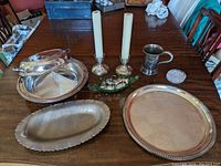 Wide shot showing all silver serving pieces on table, including divided tray with lid, oval and round trays, candlesticks, mug, green glass salt and pepper set, and small dish.