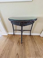 Full side view of dark wicker base half-moon small table with glass top on wooden floor and light wall background.