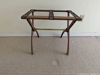 Front view of wooden suitcase stand with woven straps placed against wall on carpeted floor.