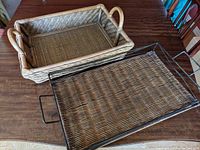 Two rectangular trays, one wicker with natural wood color and handles, one metal framed with woven base and handles, shown on wooden surface.