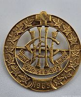 Front view of the gold pin showing the circular maple leaf border, cross symbol, CHC insignia, motto banner, and date 1965.
