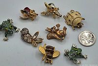 Full view of all eight animal pins including two green frogs, two bug pins, a bee pin, a squirrel pin, a ladybug pin, and a coin for size reference.