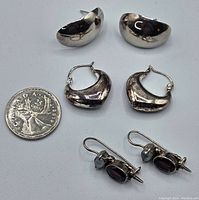 Three pairs of earrings for pierced ears displayed with a 25-cent coin for scale, showing crescent-shaped silver earrings and stone-set earrings.