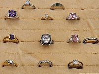 Top view of twelve assorted costume jewelry rings with various colored stones on beige velour display. The front silver-tone ring is missing a stone.