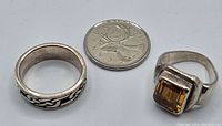Two sterling silver rings placed beside a Canadian 25 cent coin for size reference. One ring has a rectangular yellow stone with a bent shank; the other is a Celtic knot patterned band.