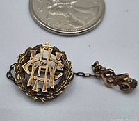 Top view of vintage 10K gold pin with chain and number 65 charm. Includes a Canadian quarter coin for size reference, showing the pin measures just under half an inch across. The pin features a detailed laurel wreath design with an intricate monogram in the center.