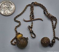 Full view of 7 inch twisted metal watch chain with attached round perfume bottle, decorative bead, T-bar and hook clasp next to a coin for scale.