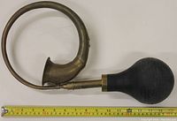 Curved brass ooga horn with black rubber bulb, showing length with measuring tape and mounting hardware