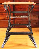 Front view of Black & Decker Workmate portable workbench showing black folding metal legs and wood slotted bench top with orange knobs for clamping.