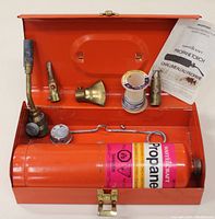 Open red metal case showing organized components of propane torch kit including tank, nozzles, Teflon tape, sparker starter, and manual.