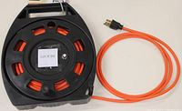 Black impact resistant cord reel with bright orange cable wrapped inside, showing power outlets and circuit breaker on one side, with the plug end of the orange cord next to it.