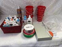 Main photo showing the variety of crafting items arranged including glue bottles in a basket, multiple red metal-handle buckets stacked, ribbon rolls, paper cutter, and plastic organizing tray.