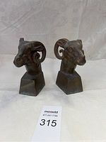 Pair of ram head brass book ends shown from front, displaying both pieces and their square bases on a white fabric background.