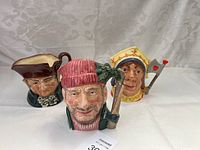Three Royal Doulton character mugs displayed side by side on a white fabric background
