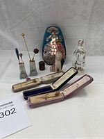 Full view of all items showing vintage hatpins, hairpins in cases, figurines including baby doll and porcelain woman, painted metal tin, and decorative containers.