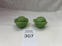 Photo of pair of green cabbage leaf salt and pepper shakers front view showing two ceramic shakers depicting textured green cabbage leaves, placed side by side on a white backdrop.