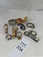 Photo showing nine watches including a gold-tone pocket watch, multiple silver and gold-tone wristwatches with various dial colors and metal bands.
