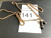 Full view of gold-tone woven chain necklace with two hand-shaped charm pendants (one black with silver accents and one silver-tone) lying flat on black surface with lot number card.