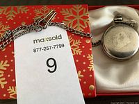 Photo of the pocket watch closed with attached chain and clip lying on white fabric and a decorative red and gold background.