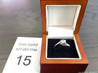 Silver-tone metal ring with clear stone displayed in open wooden box with white and black lining.