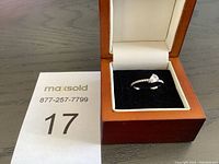 Ring displayed in open wooden box with black velvet padding, showing the full ring design and stone.