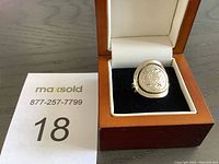 Ring displayed in wooden presentation box with black velvet insert and white interior cushioning. Focus on overall view of ring and box.