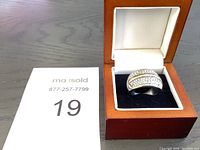 Ring displayed inside a wooden ring box with white leather interior and black velvet base, placed on a wooden surface with a MaxSold paper numbered 19 beside it.