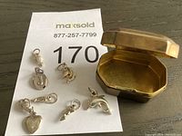 Small metal octagonal hinged box with seven silver charms arranged next to it on a wooden surface with a MaxSold lot number card underneath.