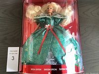 Front view of Barbie doll in original packaging wearing emerald green satin gown with decorative holly leaves and red berries on collar and waist brooch.