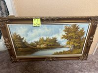 Frontal image of an oil painting depicting a cabin by a lake with trees and a partly cloudy sky in the background. The painting is housed in a large gold ornate frame with floral and scroll details at corners.