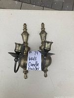 Two brass-colored wall-mounted candle holders with curved arms and single candle cups, shown on a white table.