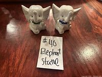 Two small white ceramic elephant statues with raised trunks, sitting on a wooden surface. One elephant has a green collar and the other a blue collar. A handwritten note states '#40 Elephant Statue'.