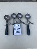 Four commercial ice cream scoops with black handles and metal scooping heads, aligned horizontally on a white surface with a note identifying them as ice cream scoops.