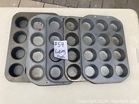 Five used metal cupcake baking trays displayed on a white surface. Trays vary in size and number of cups, with visible signs of use such as baked residue and discoloration.