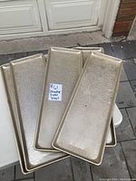 Five gold-toned decorative metal trays arranged partially overlapping on a white table outdoors, showing their textured rectangular surfaces and raised edges.