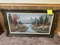 Front view of framed landscape painting depicting lake, waterfall, autumn trees, pine forest, and snow-capped mountains with ornate carved dark wood frame.