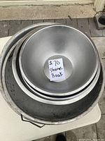 Stack of 7 large to medium metal bowls and strainers photographed from above on a white stool outdoors.