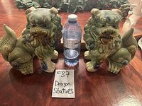 Photo showing pair of greenish-brown porcelain dragon statues on wooden surface with water bottle for scale.
